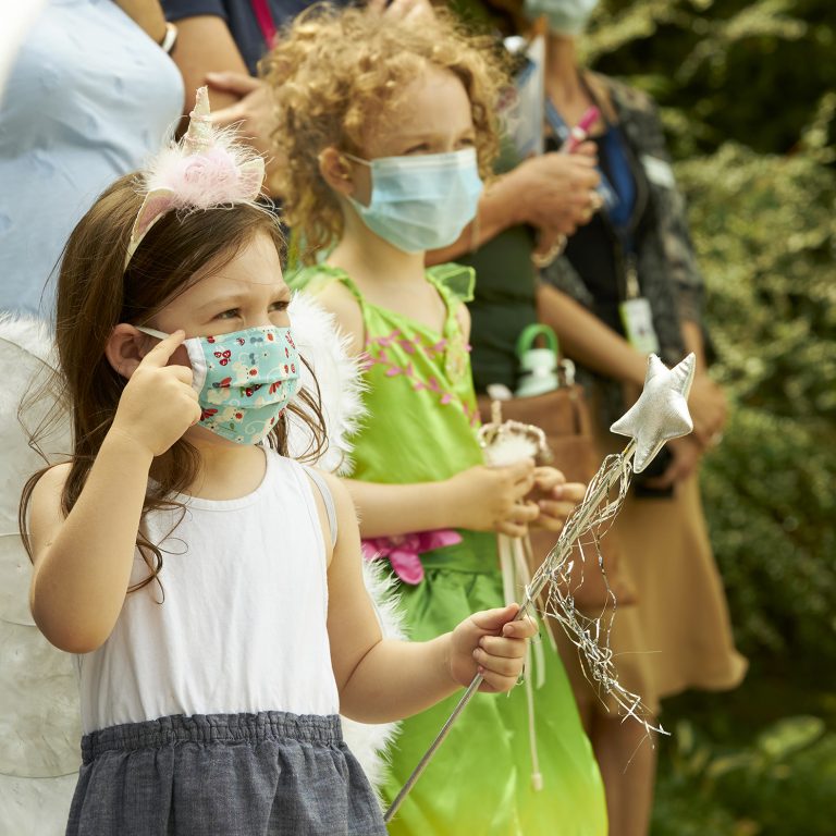 Two young girls wearing fairy wings and holding wands. They are both wearing face masks.