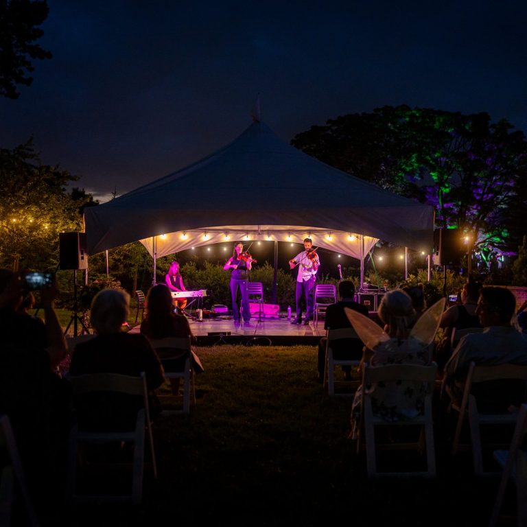 Small covered stage at rock garden with crowd watching fiddle band performing for RBG After Dark