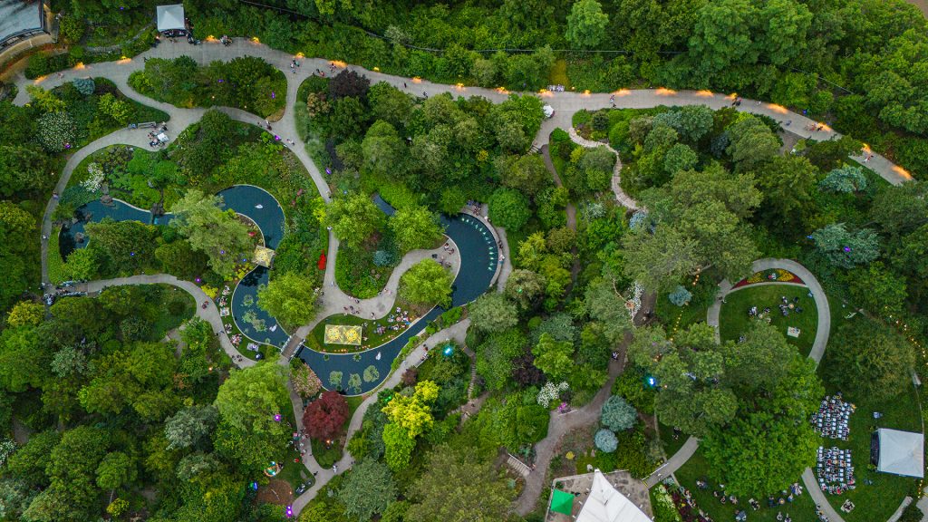 Royal Botanical Gardens RBG / July 11 2024 / Tony F Media Inc. / 10156 aerial photo of Rock Garden with a lively event below. On the left hand side, guests can be spotted relaxing in the grass and around firepits in the lower garden. On the right hand side there is a stage with rows of chairs filled with guests watching.