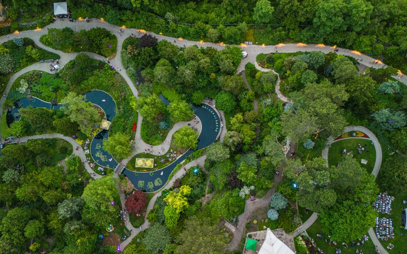 Royal Botanical Gardens RBG / July 11 2024 / Tony F Media Inc. / 10156 aerial photo of Rock Garden with a lively event below. On the left hand side, guests can be spotted relaxing in the grass and around firepits in the lower garden. On the right hand side there is a stage with rows of chairs filled with guests watching.