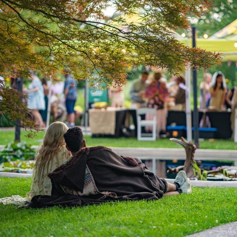 Couple reclined on the turf alongside a winding water feature in rock garden, festivities in the background