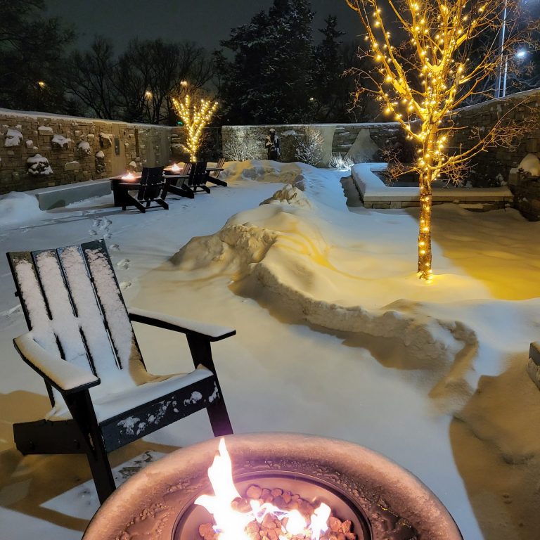 Snow covered muskoka chair behind a warm firepit. Trees in the courtyard are decorated with white string lights