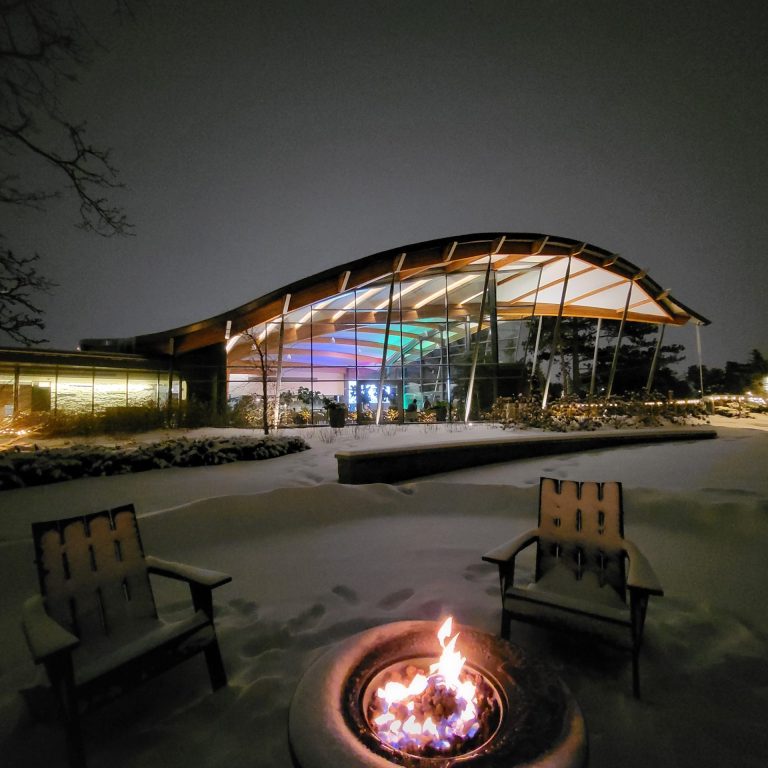 Two muskoka chairs around a warm firepit. The Rock Garden Visitor centre is lit up with coloured lights inside