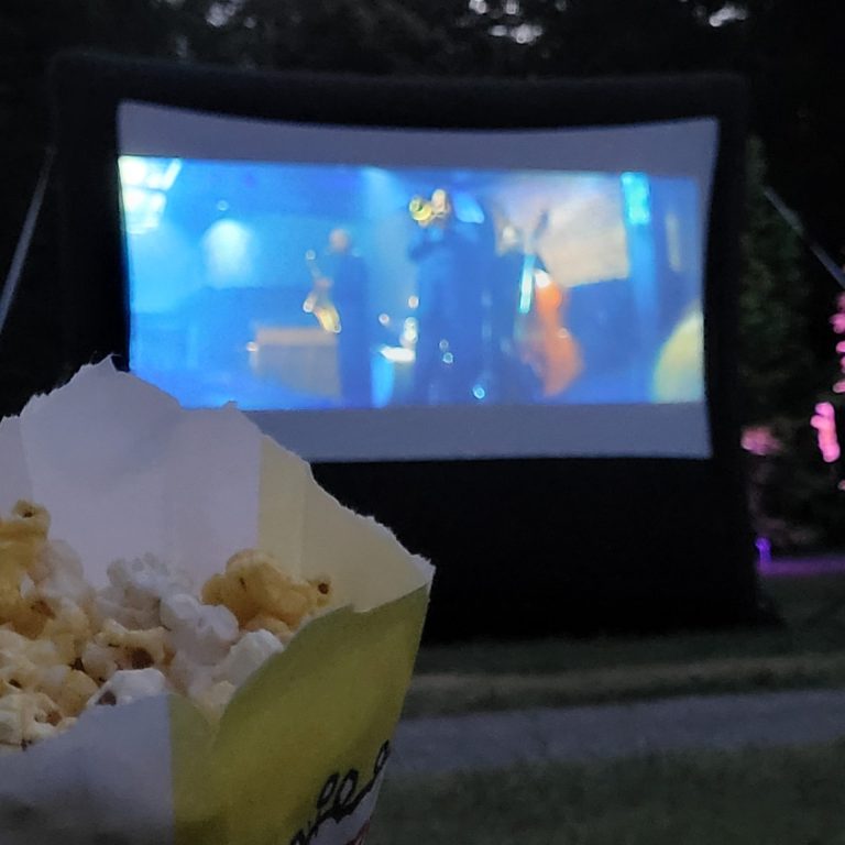 Outdoor movie screen set up in the evening with a film playing, a small bag of popcorn in the foreground