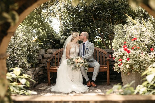 2022_Wedding_MedGarden_Geoff Shaw_01242 Bride and groom seated on a bench in a greenhouse setting in Burlington