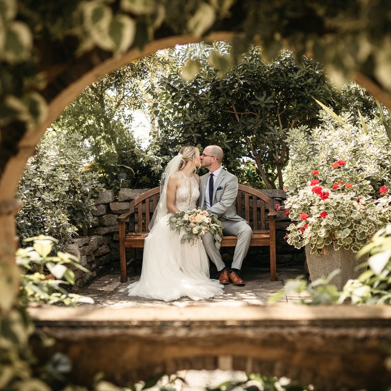 Bride and groom seated on a bench in a greenhouse setting in Burlington