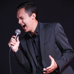 3S2A0216 Santiago Moa wearing an all-black suit singing into a microphone on a black background