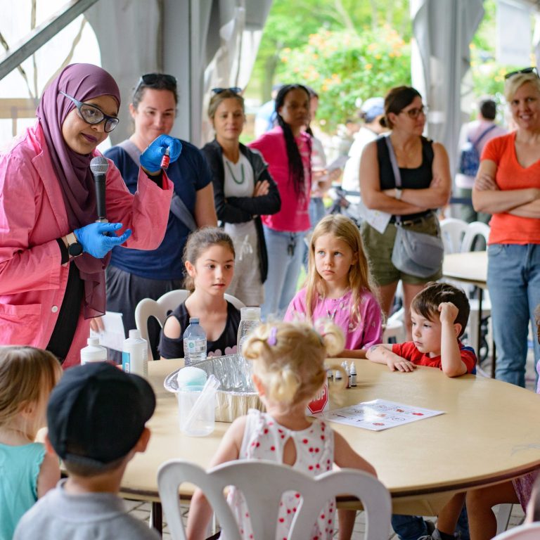 adult giving a demonstration to a group of children watching attentively. Adults stand behind them also interested.
