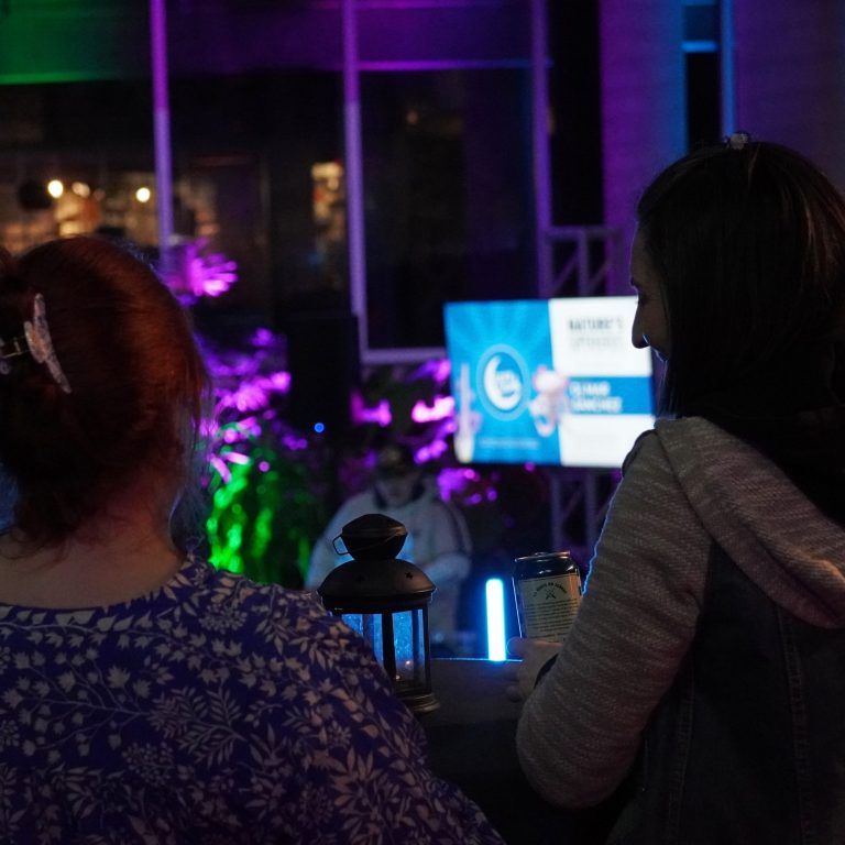 Two people at After Dark event with colorful purple and green lighting in the background. They are standing with a drink at a table. watching a DJ perform in the background.