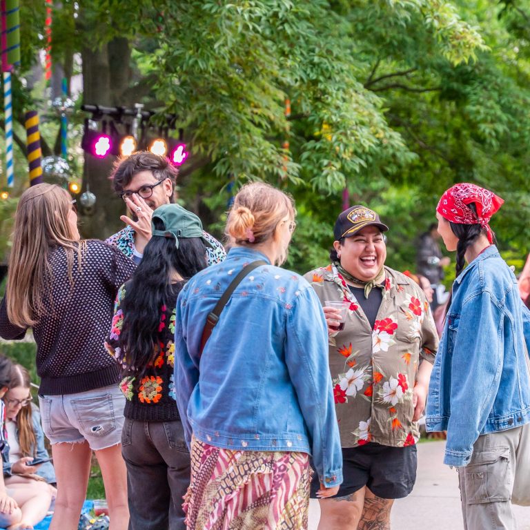 A group of people stand together outdoors in a lively and colourful garden setting, laughing and talking.