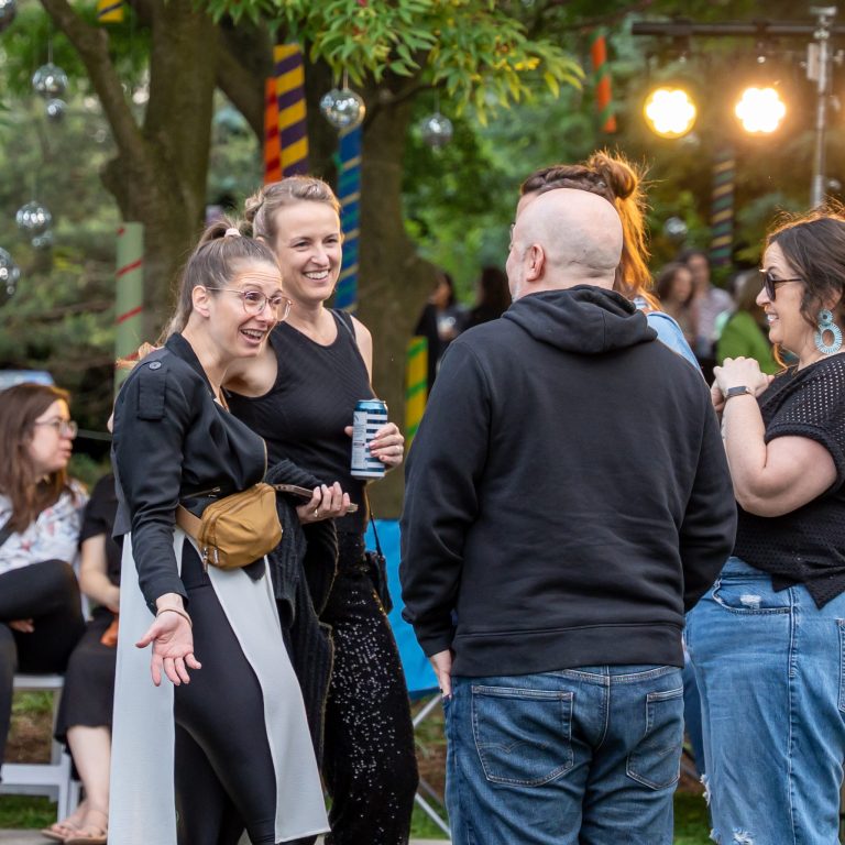 A group of people stand together outdoors in a lively and colourful garden setting, laughing and talking.