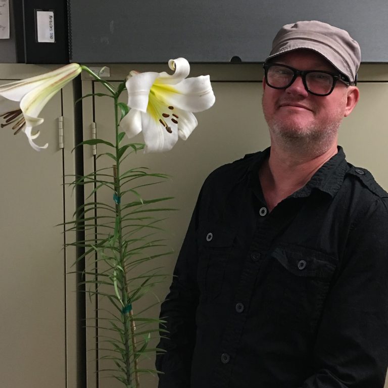 Alex Henderson standing in office with blooming Creelman Lily in a pot
