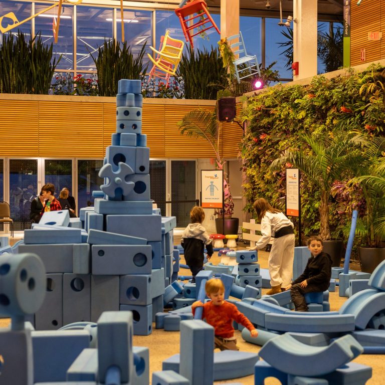 An imaginative play area with large blue foam building blocks, where children are actively constructing structures. The high ceiling showcases an artistic installation of colorful wooden chairs and keys suspended in midair, contributing to a whimsical atmosphere.