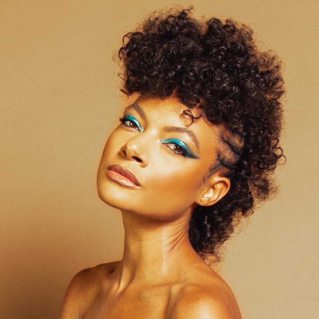 Allison Russell Allison Russell with teal eye makeup and a natural hairstyle posing on a tan background