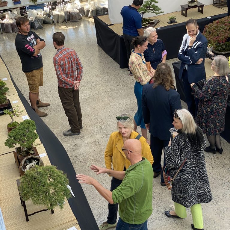 A visitor views a display of bonsai trees arranged on tables in a bright indoor exhibit space. The trees vary in size and style, with labels in front of each. Natural light streams in through tall windows behind a green living wall.
