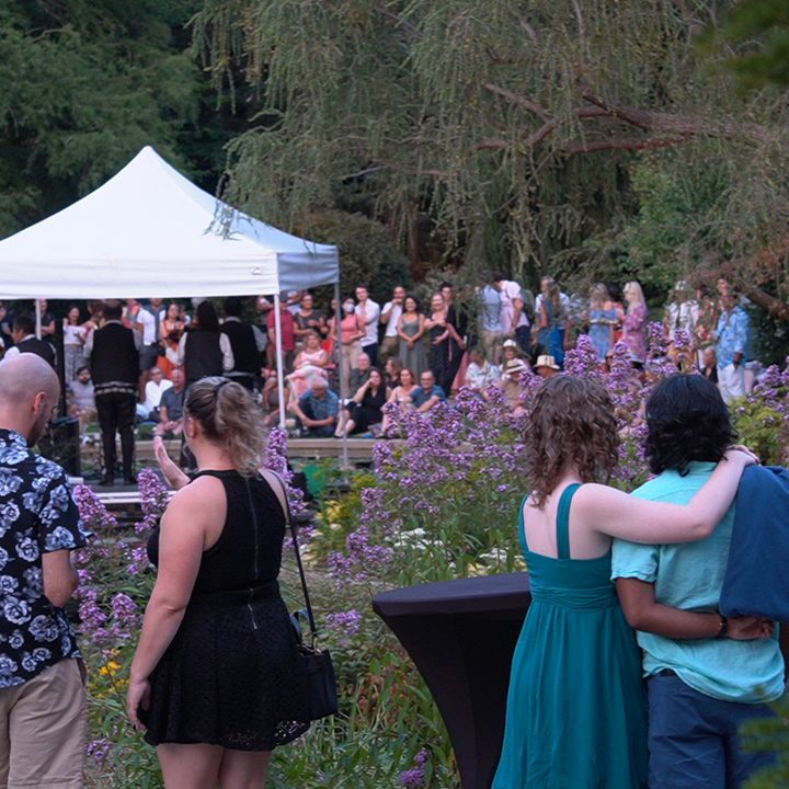 A large crowd gathers in a lush garden setting, watching a live performance under a white tent. Purple flowers bloom in the foreground, and several people stand closer to the camera, including a couple with arms around each other, enjoying the evening.