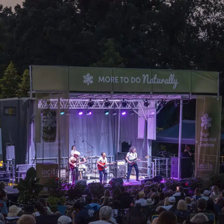 Outdoor evening concert in Hendrie Park. A band performs on a lit stage with purple lights, framed by trees and banners reading 'Royal Botanical Gardens' and 'More to Do Naturally.' A large audience watches from the foreground.