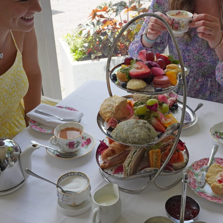 Two individuals enjoying an elegant afternoon tea in a bright, sunlit space. The table is set with a silver three-tier stand filled with colorful treats, including fresh fruit, macarons, scones, and finger sandwiches. A floral teapot, fine china cups, and saucers complete the charming setup. One person is smiling, while the other sips tea, creating a warm and inviting atmosphere.