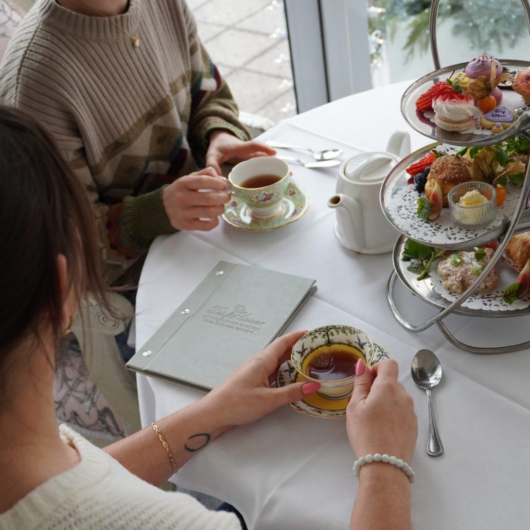 two people enjoying afternoon tea. They are each holding onto fine china cups while seated at a table with a three tiered tea platter.