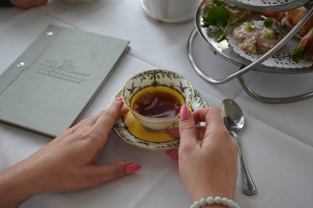 close up of hands holding a cup of tea in fine china. There is a tea platter on the table along with a Tea House menu
