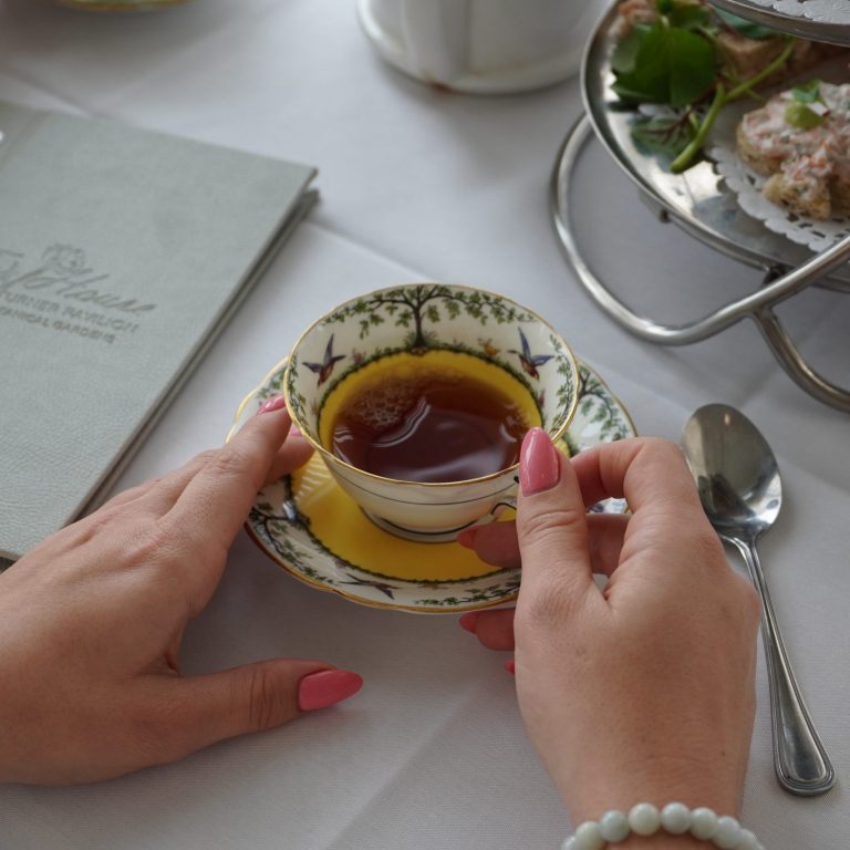 close up of hands holding a cup of tea in fine china. There is a tea platter on the table along with a Tea House menu