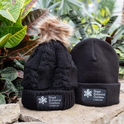A black winter toque with the Royal Botanical Gardens logo featured at the front. It's place next to a knitted RBG hat hat with a faux fur pom pom at the top and the RBG logo embroidered on the front