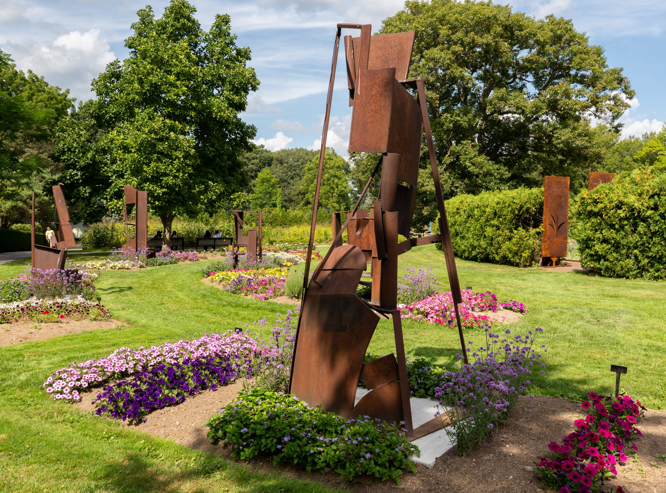 Mann Sculpture Collection - Hendrie Park - Douglas Bentham - The Hardingham Series The Hardingham Series - Tall rusted metal sculptures stand among colourful flower beds in Hendrie Park