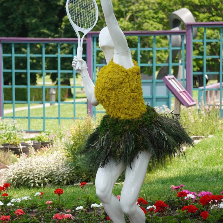 tennis player mannequin, dressed in a yellow top made of moss and a green, grass-like skirt. The sculpture is set amidst Veggie Village garden.