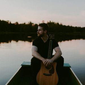 Gareth Bush Gareth Bush sitting with his guitar on a lakeside dock