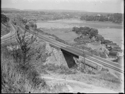 archival image of the east side of Burlington Heights in Hamilton taken from a point just south of the high level bridge across the Desjardins Canal, looking north. Pictured in the image is Carrolls Bay at right in the distance. The cluster of buildings at right in the foreground is part of the Boathouse Community, an informal subsistence settlement situated on the Burlington Heights peninsula.