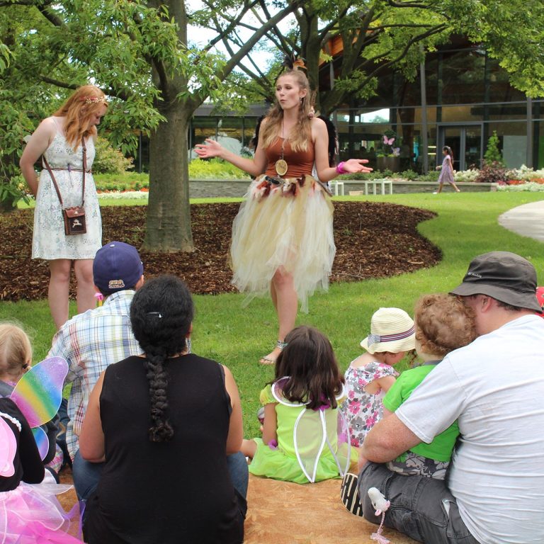 Children and parents sitting on the grass in front of two staff members dressed as fairies, telling a story