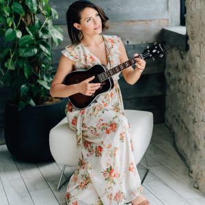 Lauren Roy Lauren Roy in floral dress sitting in a sunny, rusticly decorated corner, holding an acoustic guitar