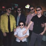 four men wearing sunglasses standing around one man sitting in a chair with sunglasses on.