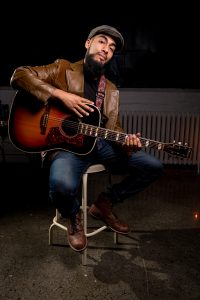 Mo Guzman sitting on a stool with a guitar