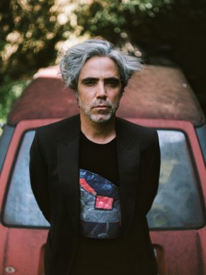 Patrick Watson leaning against an old red sedan, wearing a tshirt and blazer