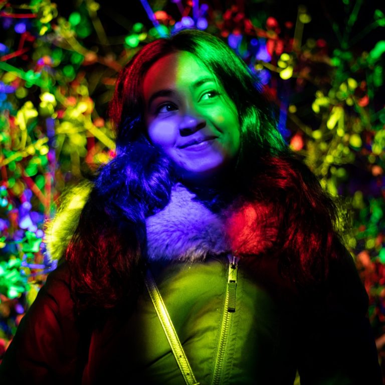 Young woman smirking and looking off to the right. Rainbow lights shine across her face