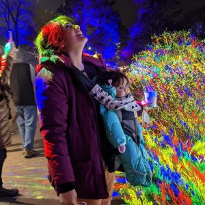 #RBGWinterWonders-MazinWakai-Rainbow-CILC Woman with baby held in front carrier looking up towards the falling snow. Rainbow lights shine across them.