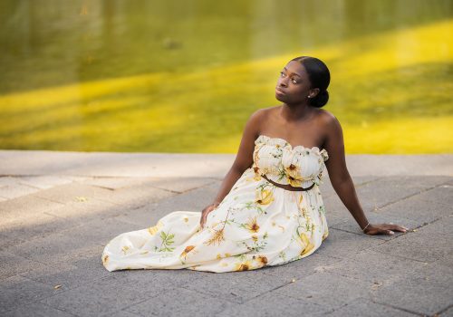 SammyJacksonHiRes1 Photo of Sammy Jackson, woman in full length, white, floral dress sits on a concrete pad, green wall in the background, in a renaissance like pose