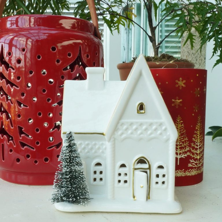 A holiday display of shop items featuring a white ceramic house with delicate gold accents, a miniature snow-dusted tree, and festive red lanterns adorned with cut-out designs and snowflake patterns.