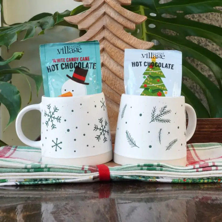 Two white holiday-themed mugs with green snowflake and pine branch designs, each holding a packet of hot chocolate mix (one with a snowman and one with a Christmas tree design). The mugs are set on a red, green, and white plaid napkin, with a wooden Christmas tree decoration and greenery in the background.