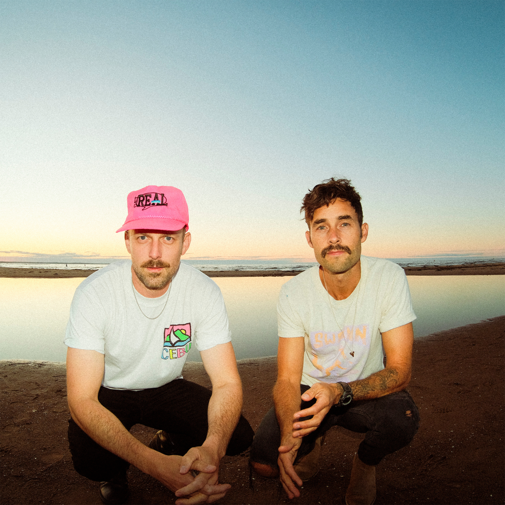 The East Pointers - Australia 2025 - 1080x1080 The East Pointers - 2 bandmates in white tshirts crouching on beach with calm waters