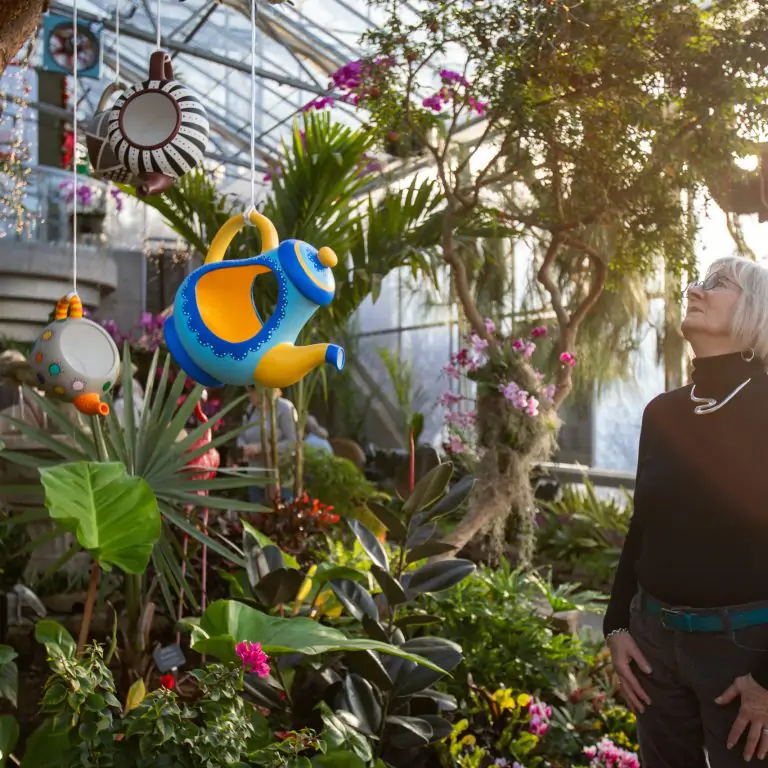 woman looking up at artistic and whimsical tea pots that are hanging from the branches of a tropical tree