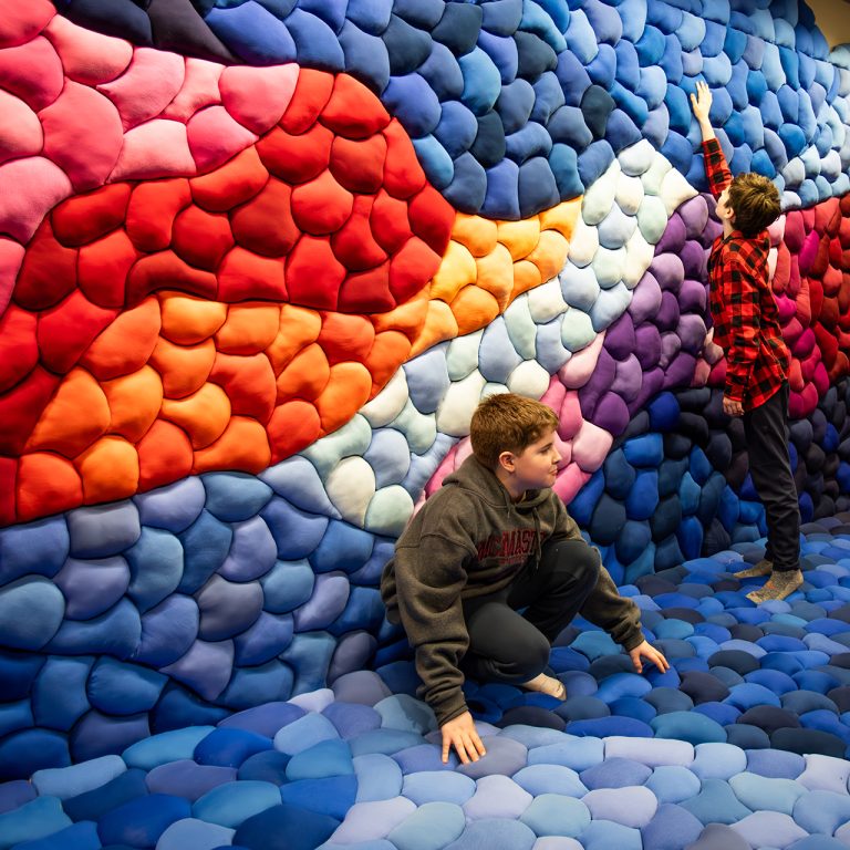 Two children interacting with a fibre arts piece of waves of stuffed colour