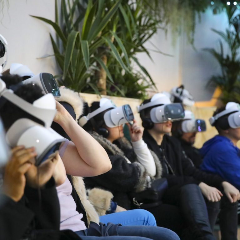 Group of people sitting in a row, wearing virtual reality headsets, immersed in a VR experience, with plants in the background creating a relaxed atmosphere.