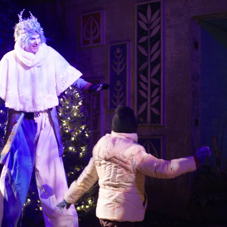 A performer dressed as a jack frost character on stilts, wearing a sparkling white cape and icy crown, interacts with a guest walking by