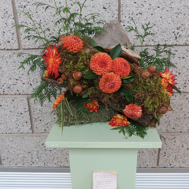 floral sculpture featuring orange dahlias, moss, wood and other organic material featured on a pedestal