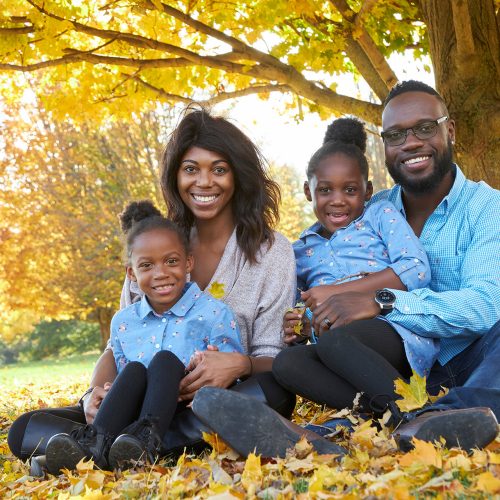 family-posing-in-autumn-arboretum Family with mother, daughter and twin daughters sitting under a tree in a pile of yellow leaves in fall