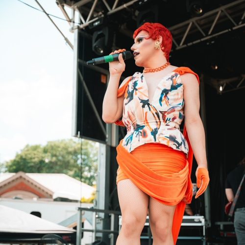 Karma Kameleon standing on stag in orange-coloured drag, speaking into a microphone