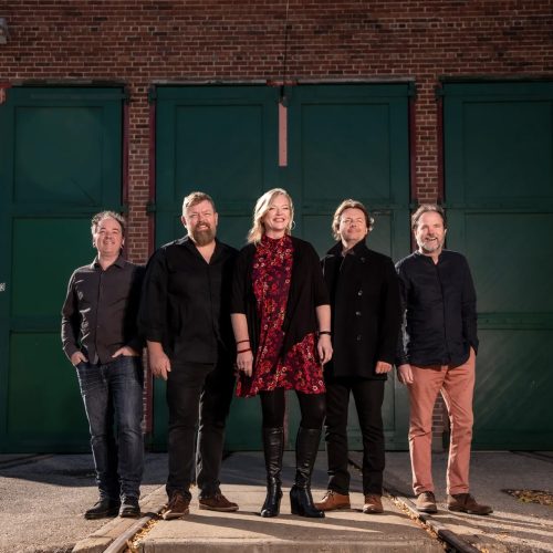 mcdades 5 band members dressed in dark warm colours stand against an industrial wall