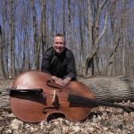 Pat sitting on a fallen tree with a large cello laying on the ground in front of him